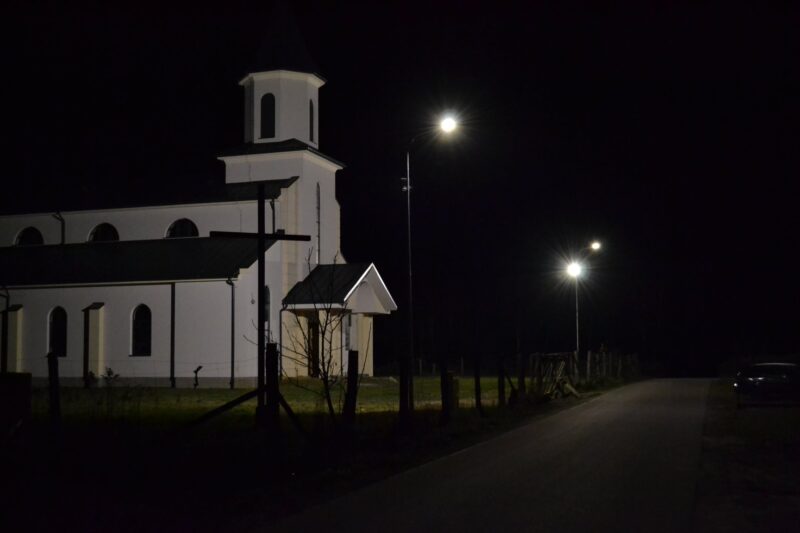 Iluminowany kosciół z wieżą i białą elewacją. Przed kościołem dwie świecące latarnie uliczne