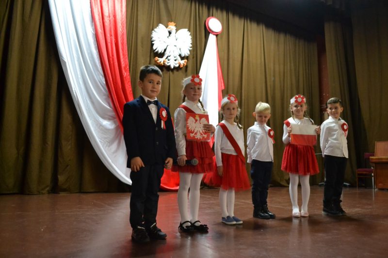 Odświętnie ubrane dzieci z biało-czerwonymi kotylionami stoja na scenie. Z tyłu sceny biało-czerwona flaga i biały orzeł z koroną