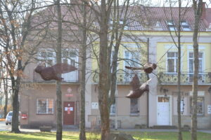 Duże ptaki wykonane z wikliny wisza między drzewami. W tle budynki