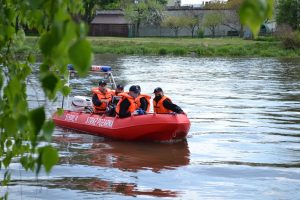 Pięciu strażaków w czarnych czapkach i pomarańczowych kamizelkach ratunkowych płynie czerowną łodzią z buałym napisem Straż Pożarna