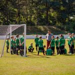 Chłopcy w zielono-czarnych strojach piłkarskich stoja na murawie obok bramki do piłki noznej. Wśród piłkarzy mężczyna w brązowych spodniach i szarej koszulce