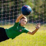 Młody bramlarz w zielonej koszulce i czarnych spodeńkach podczas obrony bramki odbija niebieska piłkę nożną