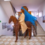 Rzeźba z wikliny - jeździec z niebieską flagą na koniu. Rzeżba ustawiona na klatce schodowej