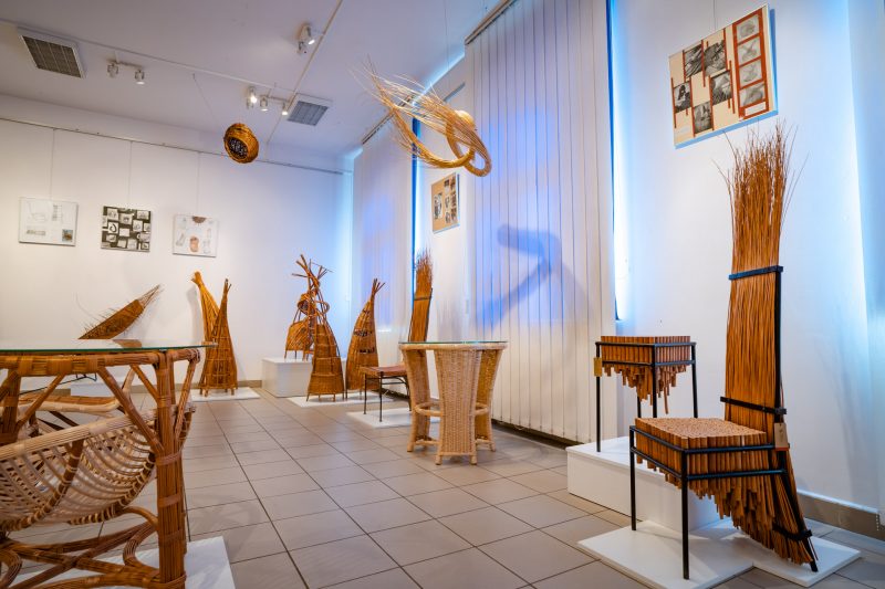 Oryginalne meble i formy z wikliny ustawiony na postumencie w sali wystawowej. Na ścianie kolorowe zdjęcia
